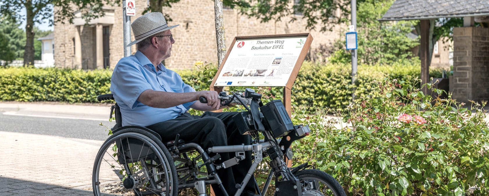Un homme en fauteuil roulant lit un panneau du sentier thématique Baukultur Eifel à Wolsfeld. En arrière-plan, on aperçoit des arbres et un bâtiment., © Naturpark Südeifel, Thomas Urbany Un homme en fauteuil roulant lit un panneau du sentier thématique Baukultur Eifel à Wolsfeld. En arrière-plan, on aperçoit des arbres et un bâtiment., © Naturpark Südeifel, Thomas Urbany
