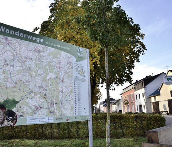 Carte de randonnée dans le parc naturel Südeifel devant des arbres et des maisons automnales à Wolsfeld. On peut également y voir un banc de repos et une boîte aux lettres., © TI Bitburger Land Carte de randonnée dans le parc naturel Südeifel devant des arbres et des maisons automnales à Wolsfeld. On peut également y voir un banc de repos et une boîte aux lettres., © TI Bitburger Land