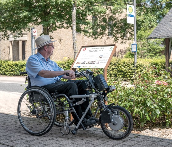 Un homme en fauteuil roulant lit un panneau du sentier thématique Baukultur Eifel à Wolsfeld. En arrière-plan, on aperçoit des arbres et un bâtiment., © Naturpark Südeifel, Thomas Urbany Un homme en fauteuil roulant lit un panneau du sentier thématique Baukultur Eifel à Wolsfeld. En arrière-plan, on aperçoit des arbres et un bâtiment., © Naturpark Südeifel, Thomas Urbany