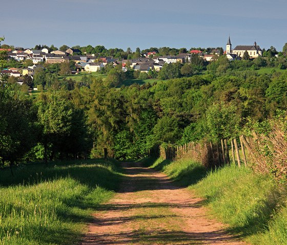 Ein Weg führt durch grüne Wiesen zu einem Dorf mit Kirche, umgeben von üppiger Vegetation unter blauem Himmel., © Naturpark Südeifel, Charly Schleder Ein Weg führt durch grüne Wiesen zu einem Dorf mit Kirche, umgeben von üppiger Vegetation unter blauem Himmel., © Naturpark Südeifel, Charly Schleder
