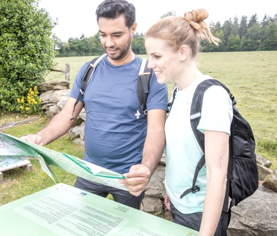 Twee mensen met rugzakken bekijken een wandelkaart voor een groene weide en bomen., © Eifel Tourismus GmbH, AR-shapefruit AG Twee mensen met rugzakken bekijken een wandelkaart voor een groene weide en bomen., © Eifel Tourismus GmbH, AR-shapefruit AG