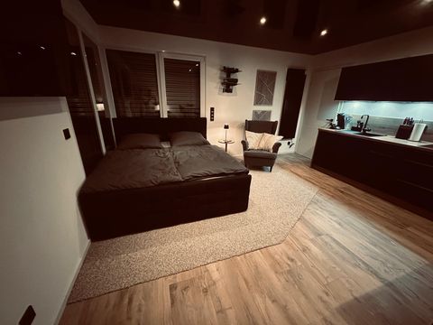 Ein modernes Schlafzimmer mit einem großen Bett, einem bequemen Sessel und einer eleganten Küchenzeile. Die Atmosphäre ist gemütlich und stilvoll, mit warmem Licht und Holzfußboden.