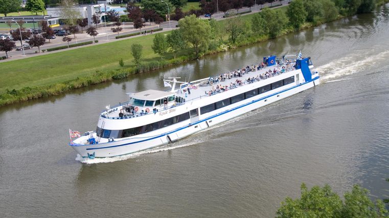 Ein großes Passagierboot fährt auf einem ruhigen Fluss. Im Hintergrund sind grüne Bäume und ein Weg zu sehen.
