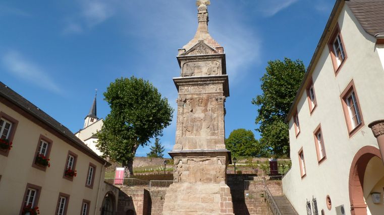 Ein imposantes Denkmal steht in einer historischen Umgebung. Umgeben von Bäumen und alten Gebäuden, bietet der Ort eine malerische Kulisse.