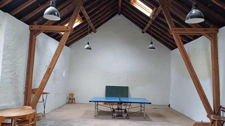 Ein heller Raum mit hohen Decken und sichtbaren Holzstützen. In der Mitte steht ein Tischtennistisch, umgeben von wenigen Stühlen.
