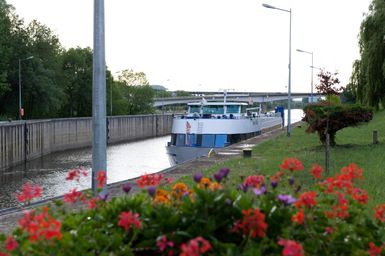 Ein Schiff fährt durch einen Kanal, umgeben von einer grünen Landschaft. Im Vordergrund blühen farbige Blumen.