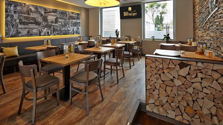 Ein modernes Restaurant mit Holzboden und gemütlichen Tischen. An der Wand ist eine große Fotografie und ein dekorativer Holzstapel zu sehen.