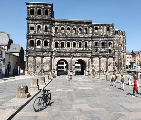 La Porta Nigra à Trèves par un temps ensoleillé. Des gens se promènent, un vélo est au premier plan. Architecture historique sur fond de ciel bleu., © TI Bitburger Land, Steffi Wagner