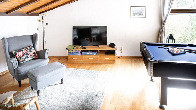 Ein modernes Wohnzimmer mit einem gemütlichen Sessel und einem Billardtisch. Der Raum hat große Fenster und eine warme Holzdecke.