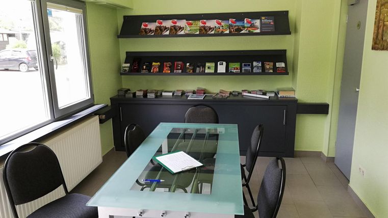 Ein einfacher Besprechungsraum mit einem gläsernen Tisch und schwarzen Stühlen. Im Hintergrund befindet sich ein Regal mit verschiedenen Broschüren.