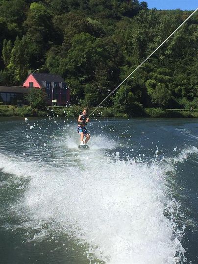 Ein Mann steht auf einem Wakeboard und wird von einem Boot über das Wasser gezogen. Im Hintergrund sind Bäume und ein pinkes Haus zu sehen.