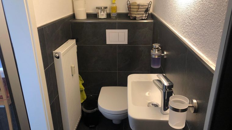 Ein kleines Badezimmer mit grau gefliesten Wänden. Es gibt ein Waschbecken, eine Toilette und einen Heizkörper.