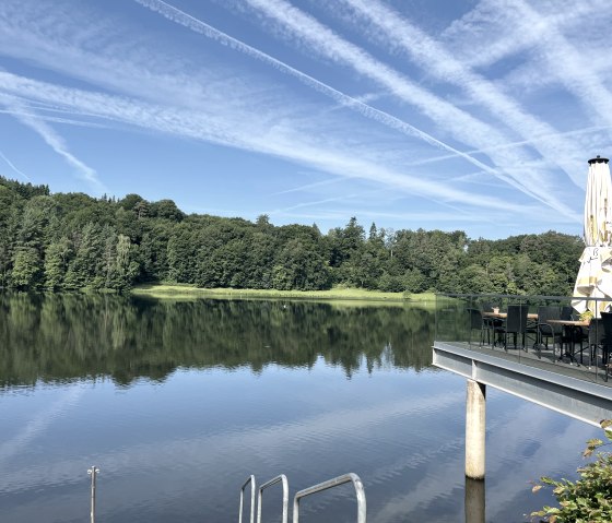 Stausee Bitburg mit Terrasse und Sonnenschirm, umgeben von Wald. Der Himmel ist blau mit Kondensstreifen., © TI Bitburger Land