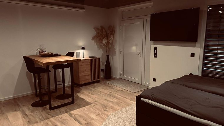 Ein modernes Zimmer mit einem Bett und einem kleinen Essbereich. Große Fenster und warme Farben schaffen eine gemütliche Atmosphäre.