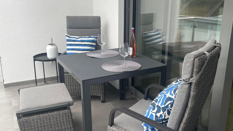 Ein gemütlicher Balkon mit einem Tisch und Stühlen aus Rattan. Auf dem Tisch stehen Gläser und eine Flasche, während ein weiteres Kissen dekorativ platziert ist.