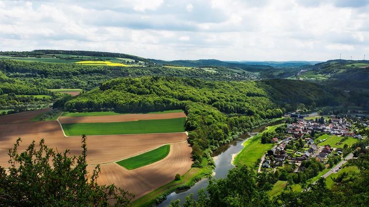 Eine malerische Landschaft mit sanften Hügeln, Feldern und einem Fluss, der durch das Tal fließt. Im Hintergrund sind Wälder und ein kleines Dorf erkennbar.