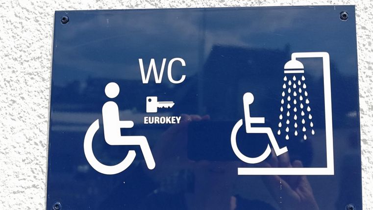 Ein Schild mit der Aufschrift "WC" und einem Eurokey-Symbol. Es zeigt Symbole für eine barrierefreie Toilette und eine Dusche.
