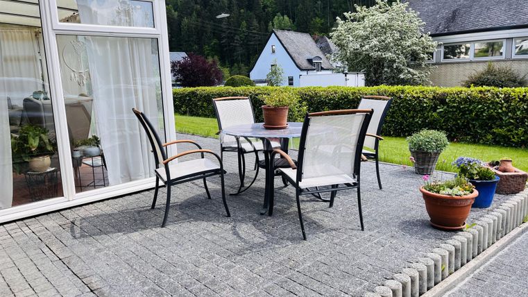 Ein gemütlicher Außenbereich mit einem Tisch und Stühlen auf einer gepflasterten Terrasse. Umgeben von schönen Pflanzen und einem grünen Rasen.