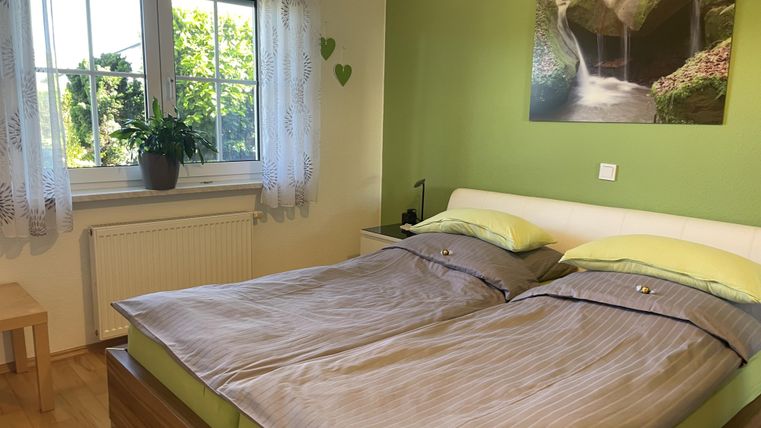 Ein gemütliches Schlafzimmer mit einem Doppelbett und grünen Wänden. Das Fenster lässt viel Tageslicht herein und wird von einem Topf mit einer Pflanze geschmückt.