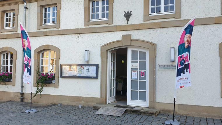 Ein historisches Gebäude mit einem einladenden Eingang. Vor dem Gebäude stehen zwei Fahnen und Blumenkästen mit Pflanzen.