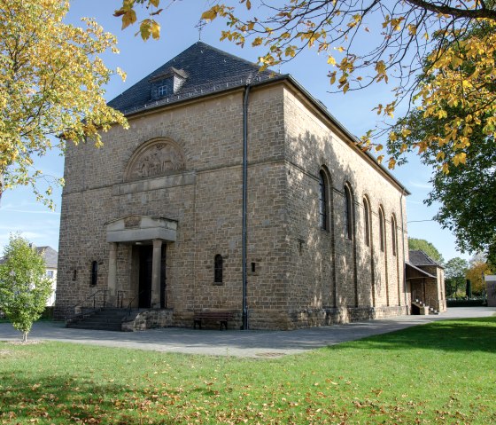 Die Kirche St. Hubertus in Wolsfeld, umgeben von herbstlichen B&auml;umen, steht in einem gr&uuml;nen Park. Das Geb&auml;ude ist aus Stein mit einem Satteldach., &copy; TI Bitburger Land