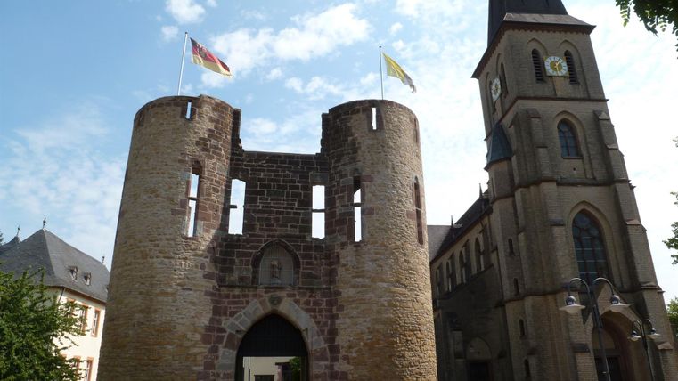 Eine historische Stadtmauer mit zwei Türmen und Fahnen. Daneben steht eine beeindruckende Kirche mit einem hohen Turm.