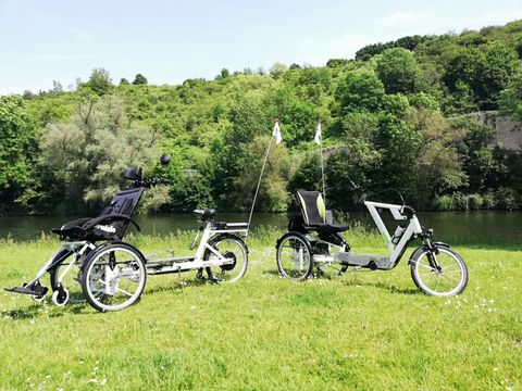 Zwei Liegedreiräder stehen auf einer grünen Wiese neben einem Fluss. Im Hintergrund sind Bäume und Hügel zu sehen, bei klarem Himmel.