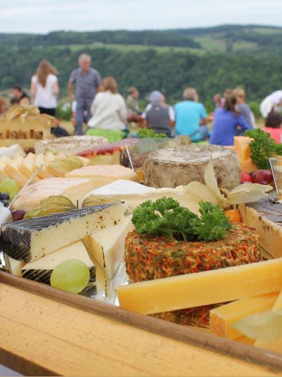 Ein reichhaltiges Käsebrett mit verschiedenen Käsesorten und frischen Trauben. Im Hintergrund sind viele Menschen in einer malerischen Landschaft zu sehen.