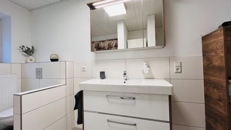 Ein modernes Badezimmer mit einem schlichten Waschbecken und Spiegel. Die Wände sind hell gefliest, und es gibt eine Holzkommode auf der rechten Seite.