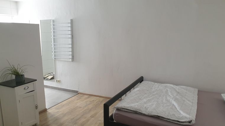 Ein einfaches Schlafzimmer mit einem Bett und einem kleinen Tisch. Die Wände sind weiß und der Boden ist aus Holz.