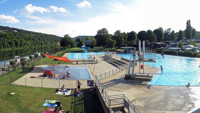 Ein Schwimmbad mit mehreren Becken und vielen Besuchern. Die Umgebung ist grün und es gibt Spielgeräte für Kinder.
