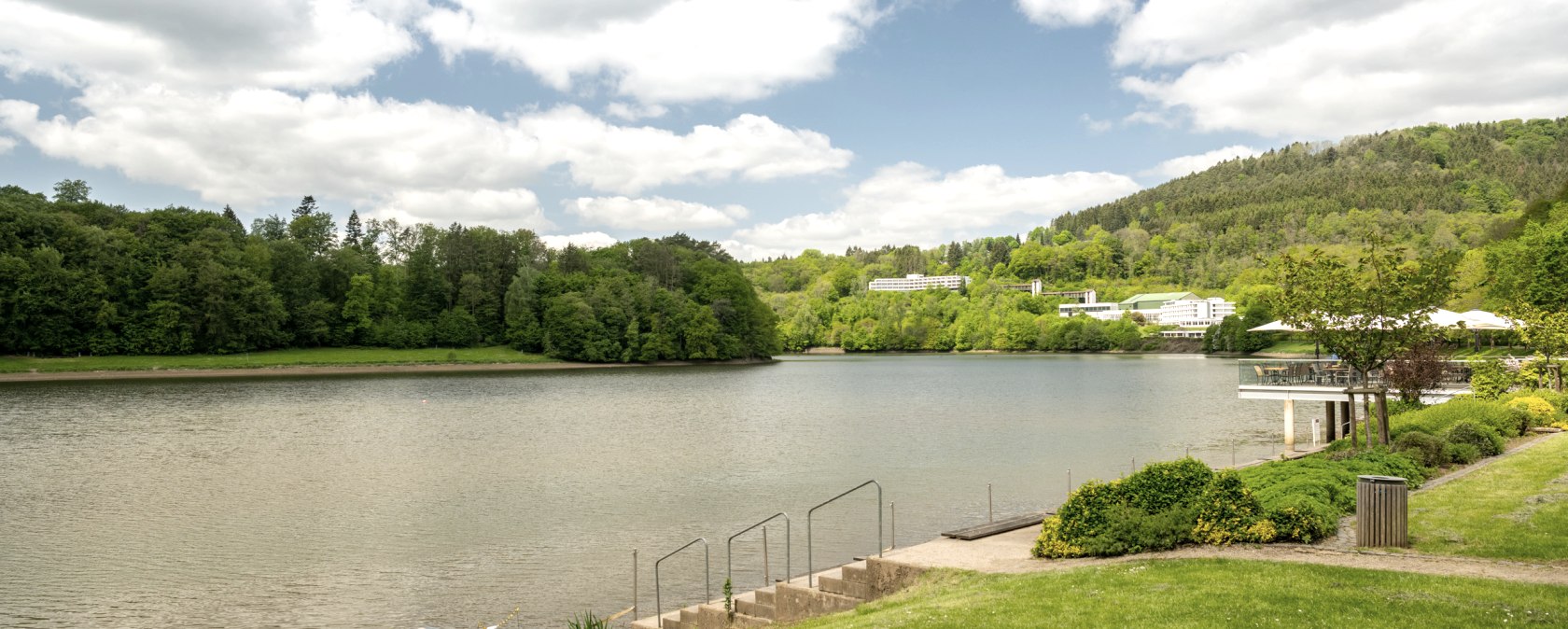 Stuwmeer van Bitburg met groen oeverlandschap, bomen en gebouwen op de achtergrond onder een bewolkte hemel., &copy; Eifel Tourismus GmbH, Dominik Ketz
