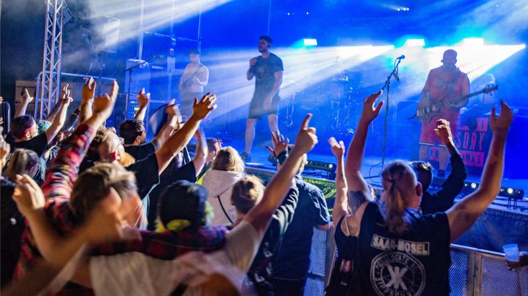 A large crowd is cheering at a live concert. The stage is illuminated, and the artists are performing with energy.