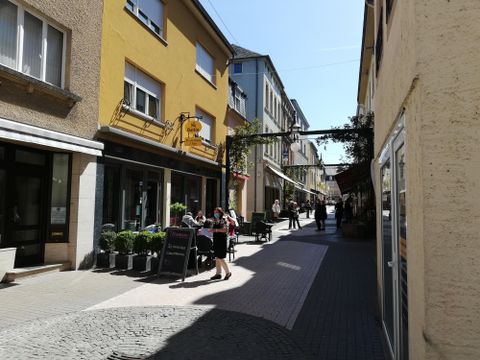 Eine charmante Straße mit bunten Gebäuden und Terrassen. Menschen sitzen gemütlich und genießen das sonnige Wetter.