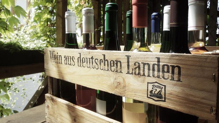 Ein Holzkasten mit verschiedenen Weinen aus deutschen Landen. Im Hintergrund sind grüne Pflanzen zu sehen.