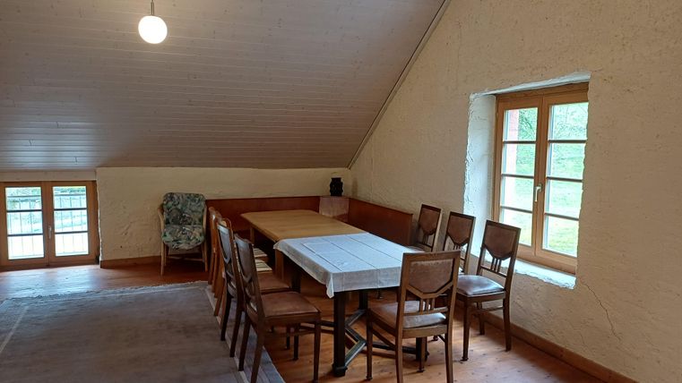 Ein gemütlicher Raum mit einem großen Tisch und Stühlen. Es gibt Fenster, die viel Licht hereinlassen, und eine kühle, einladende Atmosphäre.