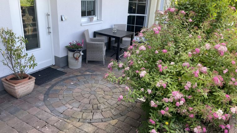 Ein gemütlicher Außenbereich mit einem kleinen Tisch und Stühlen. Bunte Blumen und ein gepflasterter Boden ergänzen den ruhigen Platz.