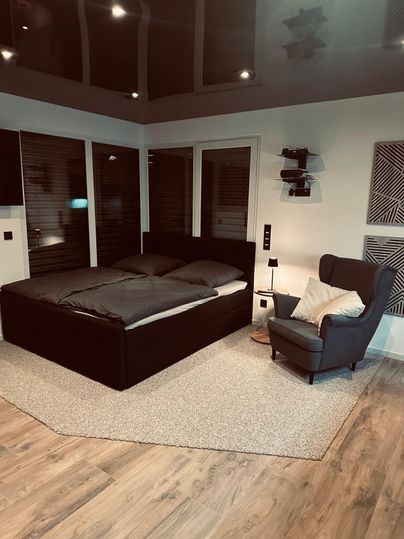 Ein modernes Schlafzimmer mit einem großen Doppelbett und einem gemütlichen Sessel. Der Raum hat einen warmen Holzboden und große Fenster, die für viel Licht sorgen.