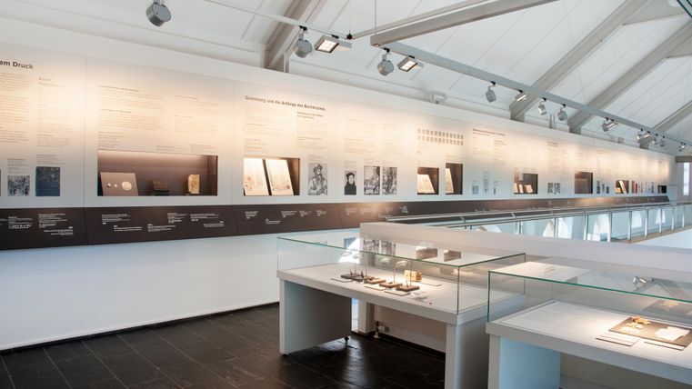 Een modern museum met een wand vol informatie en tentoonstellingen. Op de voorgrond zijn vitrines met verschillende objecten te zien.