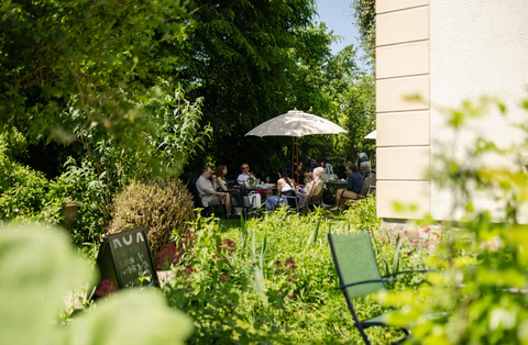 Ein sonniger Garten mit Gästen, die unter Sonnenschirmen sitzen. Umgeben von vielen Pflanzen und einer einladenden Atmosphäre.