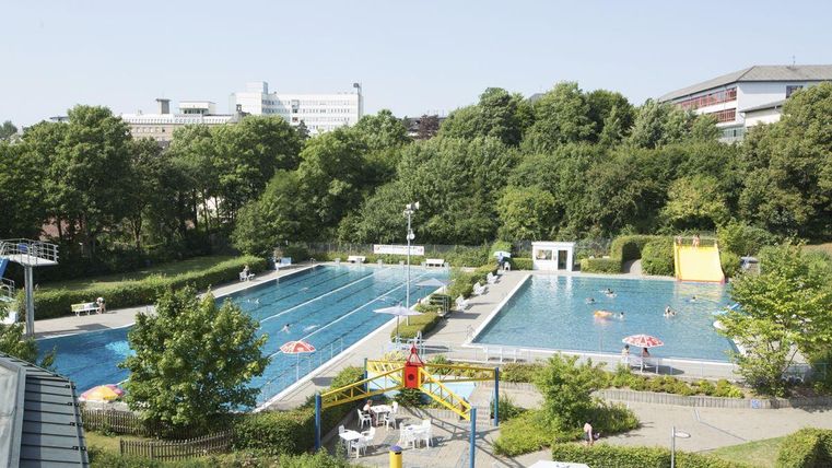 Ein Freibad mit mehreren Schwimmbecken und grünen Bäumen im Hintergrund. Sonnenschirme und Badegäste verleihen der Szene eine sommerliche Atmosphäre.
