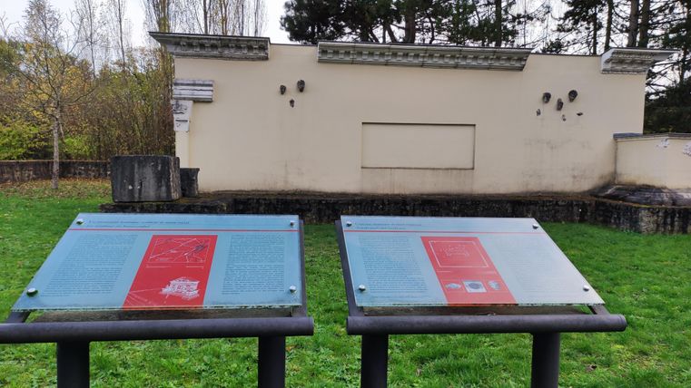 Zwei Erklärtafeln stehen vor einer alten Wand in einer parkähnlichen Umgebung. Im Hintergrund sind Bäume zu sehen, und das Gelände ist mit grünem Gras bedeckt.