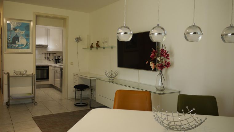 Ein modernes Wohnzimmer mit einem großen TV, einem Esstisch und bunten Stühlen. Im Hintergrund ist die Küche sichtbar, die hell und einladend wirkt.