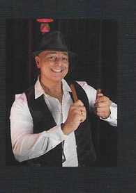 Een man in een hoed met een glimlach, die met een hand aan een object werkt. Op de achtergrond is een zwarte gordijn te zien.