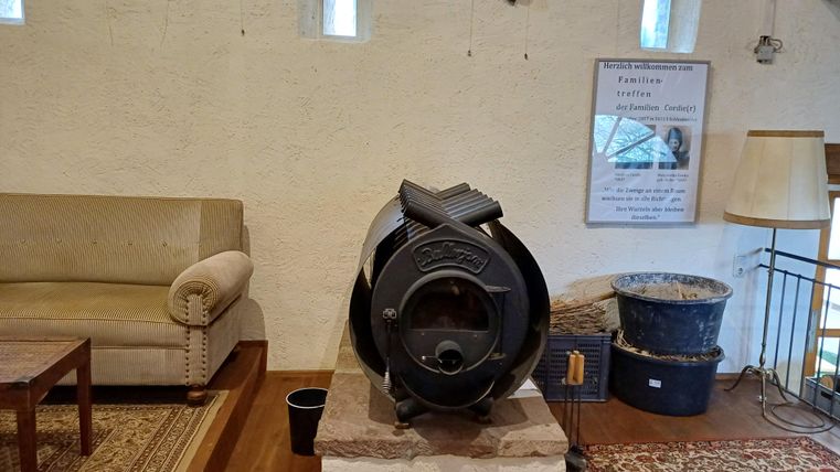 Ein gemütlicher Raum mit einem Holzofen auf einem Steinpodest. Dahinter stehen ein Sofa, ein Tisch und weitere Möbel.