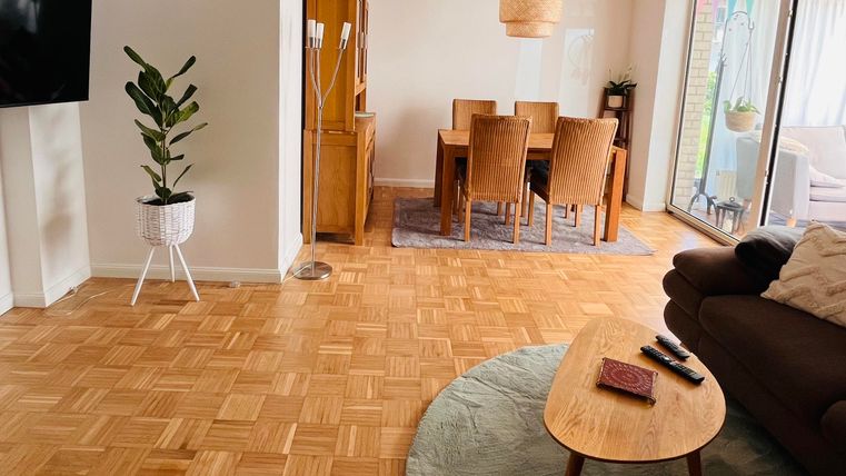 Ein modernes Wohnzimmer mit einem Holzfußboden, einer gemütlichen Couch und einem runden Tisch. Im Hintergrund steht ein Essbereich mit einem großen Tisch und Pflanzen.