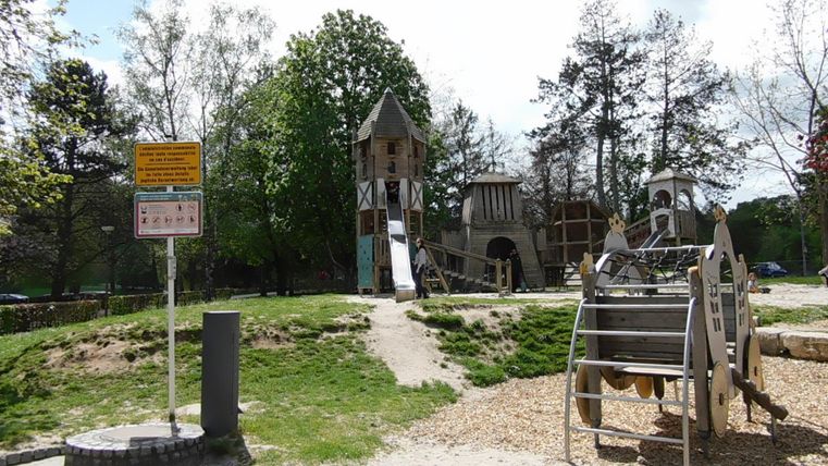 Ein Spielplatz mit einem großen Spielturm und verschiedenen Spielgeräten. Umgeben von Bäumen und Grünflächen bietet er eine einladende Spielumgebung.