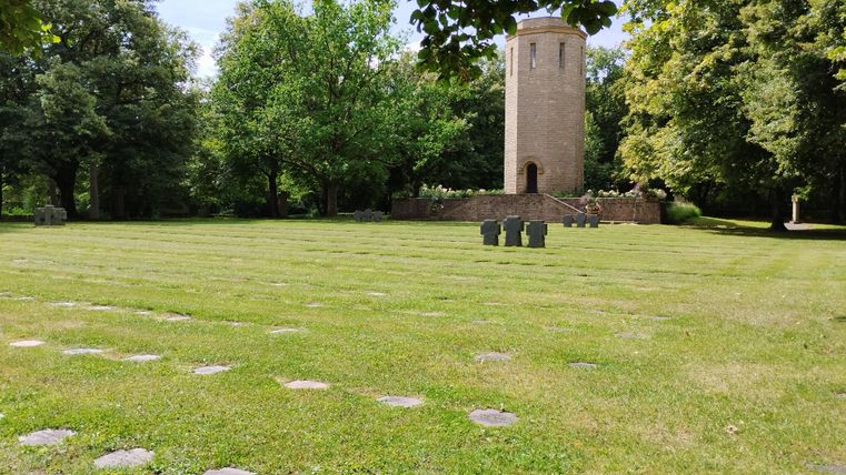 Ein grüner Park mit einem runden Turm im Hintergrund. Im Vordergrund sind einige Steine auf dem Rasen verteilt.