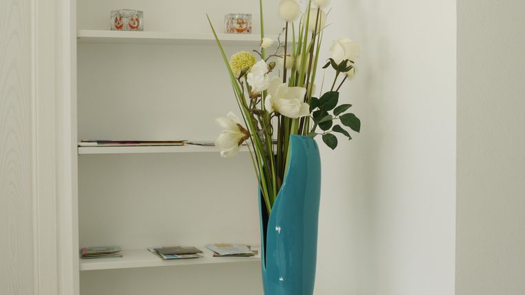 Eine moderne Vase in leuchtendem Blau mit weißen Blumen steht auf einem gläsernen Tisch. Im Hintergrund sind Regale mit verschiedenen Heften und Objekten sichtbar.