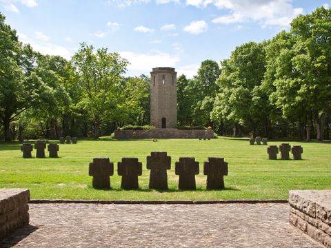 Kriegsdenkmal mit Steinkreuzen und Turm im Hintergrund, umgeben von Bäumen.